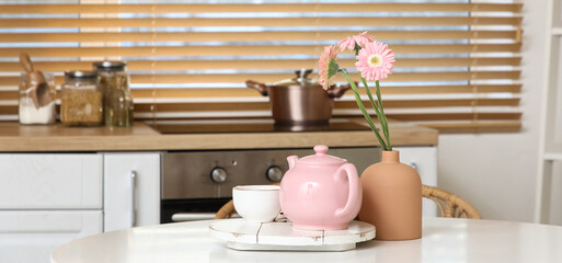 Pink teapot with cup of tea and beautiful flowers on table in kitchen