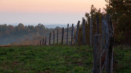 Barbed wire fence curving over hill during dusk, rural landscape with autumn colors on forest and orange glow in sly
