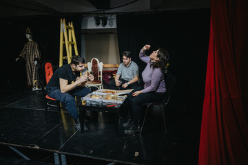 Performers gather on theater stage, eating and socializing during a break, surrounded by costumes and equipment, highlighting the camaraderie and creative atmosphere of professional stage productions.