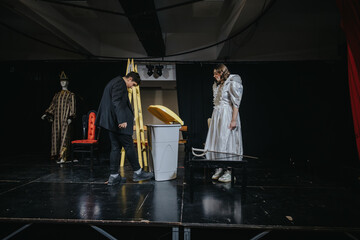 Two young actors rehearse a scene on stage with theatrical props and costumes, surrounded by a...