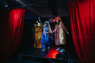 A group of performers dressed in vibrant costumes act on a dimly lit stage with red curtains,...