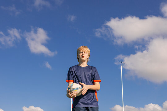 Junior rugby union player standing on field holding ball with goal posts in background