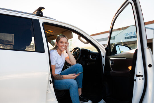 First Nations Australian woman sitting in drivers seat of car with door open scrolling mobile phone