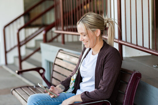 young woman seated on park bench chair in urban area of country town with phone