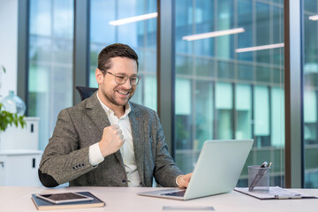 Happy young businessman sitting at a desk in the office, looking at the laptop screen and rejoicing at the news and smiling, showing a victory gesture with his hand