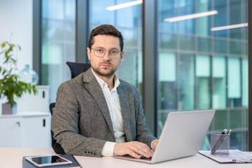 Fototapeta premium Portrait of a serious and confident young man in a suit and glasses sitting in the office at a desk, working on a laptop and looking at the camera