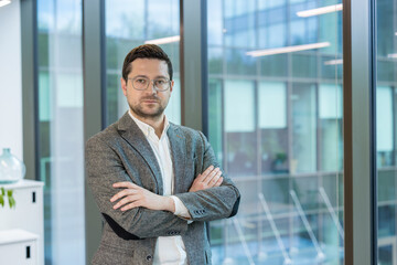 Portrait of a serious young businessman in a suit and glasses standing in the office by the window,...