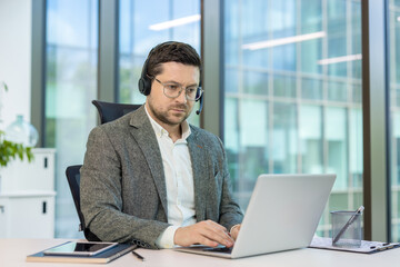 Serious young man in a suit and headset working intently on a laptop while sitting at a desk in the...