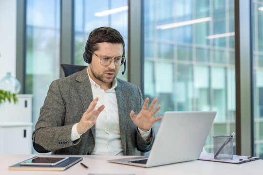 Confused young male businessman in headset and suit working at laptop, talking worriedly on video call, gesturing with hands