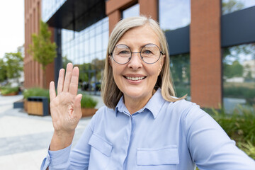 Smiling senior woman waves during a video call, possibly using a phone, outdoors near a modern...