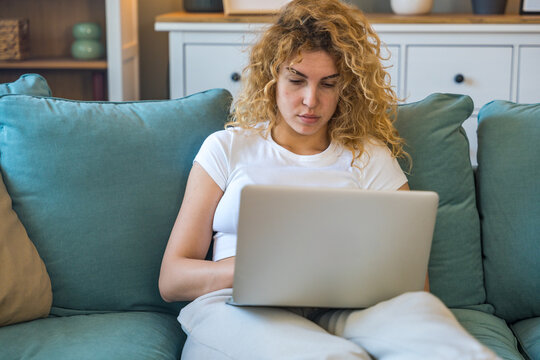 Young university student working side hustle to pay rent and costs of living as translator. Woman translating text on her laptop computer while sitting on sofa at home. Female typing online message. - Powered by Adobe