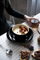 Bowl of semolina porridge with walnuts, red currants and apples on a black plate. Woman’s hand with tea, spoon, and toast on cozy morning breakfast table