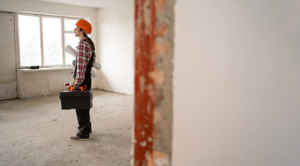 Builder, construction worker or repairman in blue overalls and protective helmet holding toolbox standing in old apartment ready for renovation