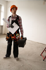male worker inside flat. Builder in hardhat and work uniform overalls standing in empty room holding toolbox, clipboard and blueprints. Repairs, constructions and renovations