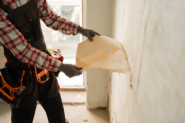 the hands of a master removing old wallpaper from a wall in an apartment before renovation