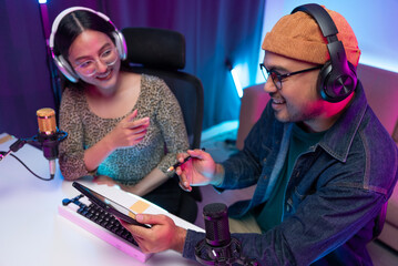 Two young asian streamer host making live podcast with condenser microphone and tablet at broadcast...