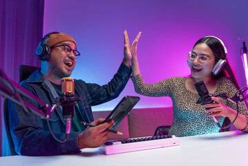 Two young asian streamer host making live podcast with condenser microphone and tablet at broadcast...