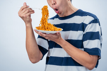 Asian man in striped shirt lifting a fork full of spicy instant noodles from a bowl while smiling...