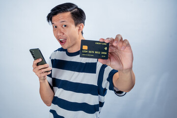Asian man in striped shirt holding a smartphone in one hand and showing a credit card toward the camera with a surprised expression, against a clean studio background