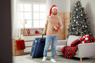 Young man with suitcase and gift box in living room. Christmas travel concept