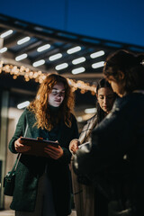 Three individuals gathered outside at night beneath decorative lights, engaged in collaborative discussion and planning using a clipboard, fostering teamwork and communication.