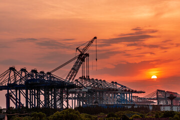 The silhouette of large lifting machinery at the seaside shipping terminal during sunset at dusk.