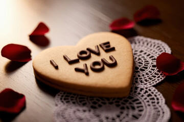 Heart-shaped cookie with "i love you" message surrounded by red rose petals on lace doily