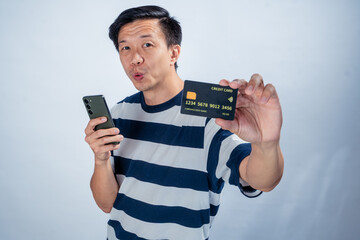 Asian man in striped shirt holding a smartphone in one hand and showing a credit card toward the camera with a surprised expression, against a clean studio background