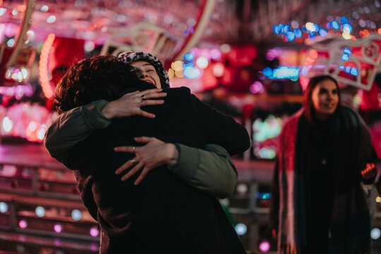 A heartfelt moment as two friends share a hug amidst a colorful carnival setting. One stands nearby in a cheerful and lively winter evening atmosphere, evoking themes of friendship and celebration.
