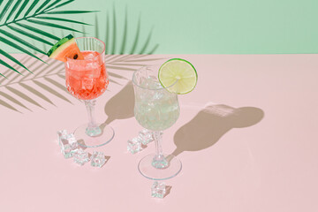 Summer scene with refreshing watermelon and lime drinks and palm leaves shadow on pink and green background. Sun and shadows. Minimal cocktail concept. Trendy summertime party idea. Summer aesthetic.