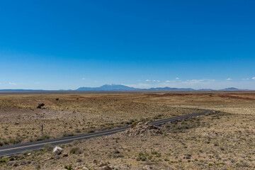 A winding desert road cuts through the vast, barren landscape near Meteor Crater in Winslow, Arizona, with distant mountains under a clear blue sky.