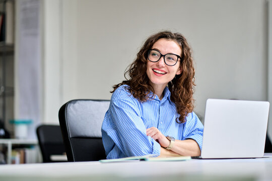 Young happy business woman company employee sitting at desk working on laptop. Smiling female worker marketer using computer in corporate modern office looking away advertising professional services.