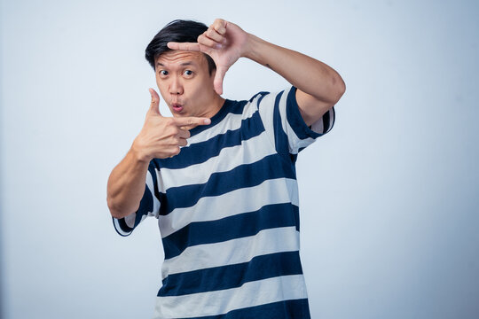 Asian man in striped shirt playfully framing his eye with fingers, pretending to take a photo, smiling against a plain background
