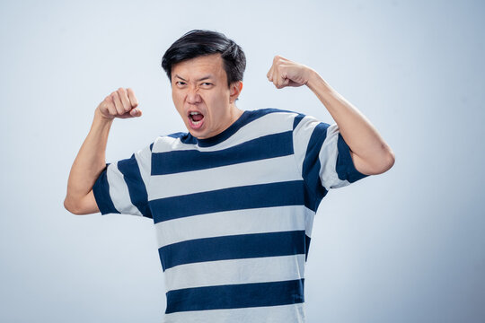Asian man in a striped shirt flexing both arms and shouting with an intense facial expression, conveying strength or anger, against a plain light background