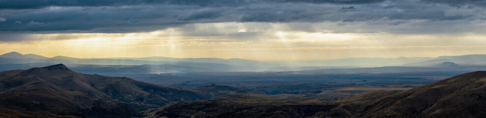 Rolling hills and distant mountains lie under a stormy sky. Iluminating flat-topped rock stands out. Neuquen arid landscape, Argentina