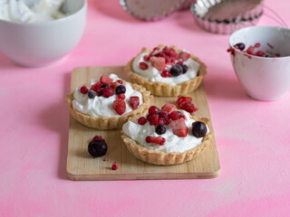 Handmade yogurt cream and red fruit tartlets on a light wooden board and with ingredients and kitchen utensils. Pastel pink background