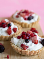 Three refreshing and healthy yogurt cream and red fruit tartlets on a light wooden board and pastel pink background