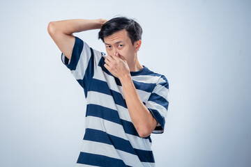 Asian man in a striped shirt holding his nose with a disgusted expression while sniffing under his...