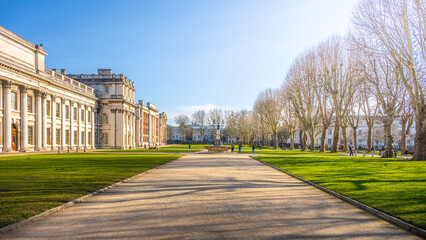 Naklejka premium Visitors stroll through the well-maintained gardens of the Old Royal Naval College in Greenwich, enjoying the sunny day and historic architecture framed by leafless trees.
