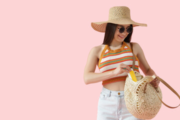 Young woman taking sunscreen cream from beach bag on pink background
