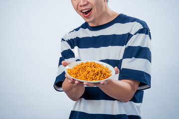 Asian man in striped shirt excitedly presenting a plate of spicy instant noodles toward the camera with a surprised facial expression against a clean white background