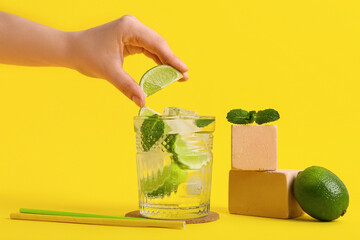 Female hand squeezing lime into glass of fresh Mojito cocktail on yellow background