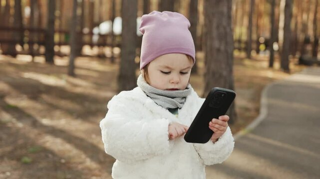 A child holds a smartphone, looks at the screen, taps it, takes pictures with a concentrated expression. A kid in a park in the fall. The child is taking pictures of nature. A little blogger