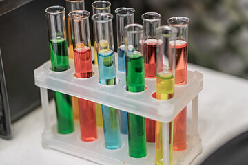 White plastic test tube rack containing nine colored liquids, lab setting with gray surface and...