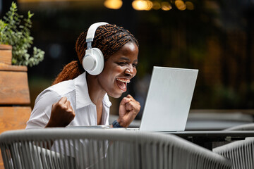 Close up view of young African woman sitting in cafe and working on laptop computer. Holding fists and celebrating success.