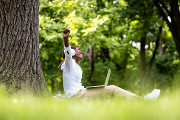 Beautiful African female freelancer working on laptop computer in nature. Raising her arms for success.
