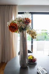 A metallic vase with daisies and orchids on a table with fruit and cutlery. A glass door opens to a balcony with greenery and natural light.