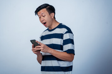 Fototapeta premium Asian man in a striped shirt holding a smartphone with a surprised and playful expression, looking directly at the camera against a plain background