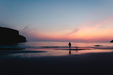 Fototapeta premium Silhouetted person and dog standing on Cornwall beach at sunset with ocean and pink sky in summer, Cornish Coastline, Mawgan Porth, England, United Kingdom, UK