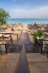 A wooden pathway leads to a sandy beach lined with thatched umbrellas and sun loungers. The turquoise sea and lush greenery create a tropical setting.